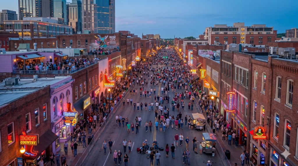 Lower Broadway in Nashville at dusk with neon honky-tonk signs, rooftop bars, and crowds on the pedestrian strip.